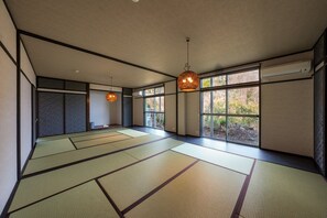 Une pièce avec un sol recouvert de tatamis, de grandes fenêtres, un ventilateur de plafond et des lampes suspendues.