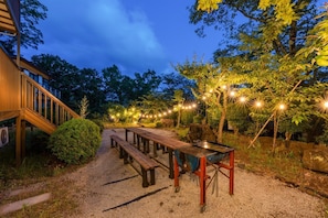 A wooden deck with benches and a barbecue grill, surrounded by trees and string lights.