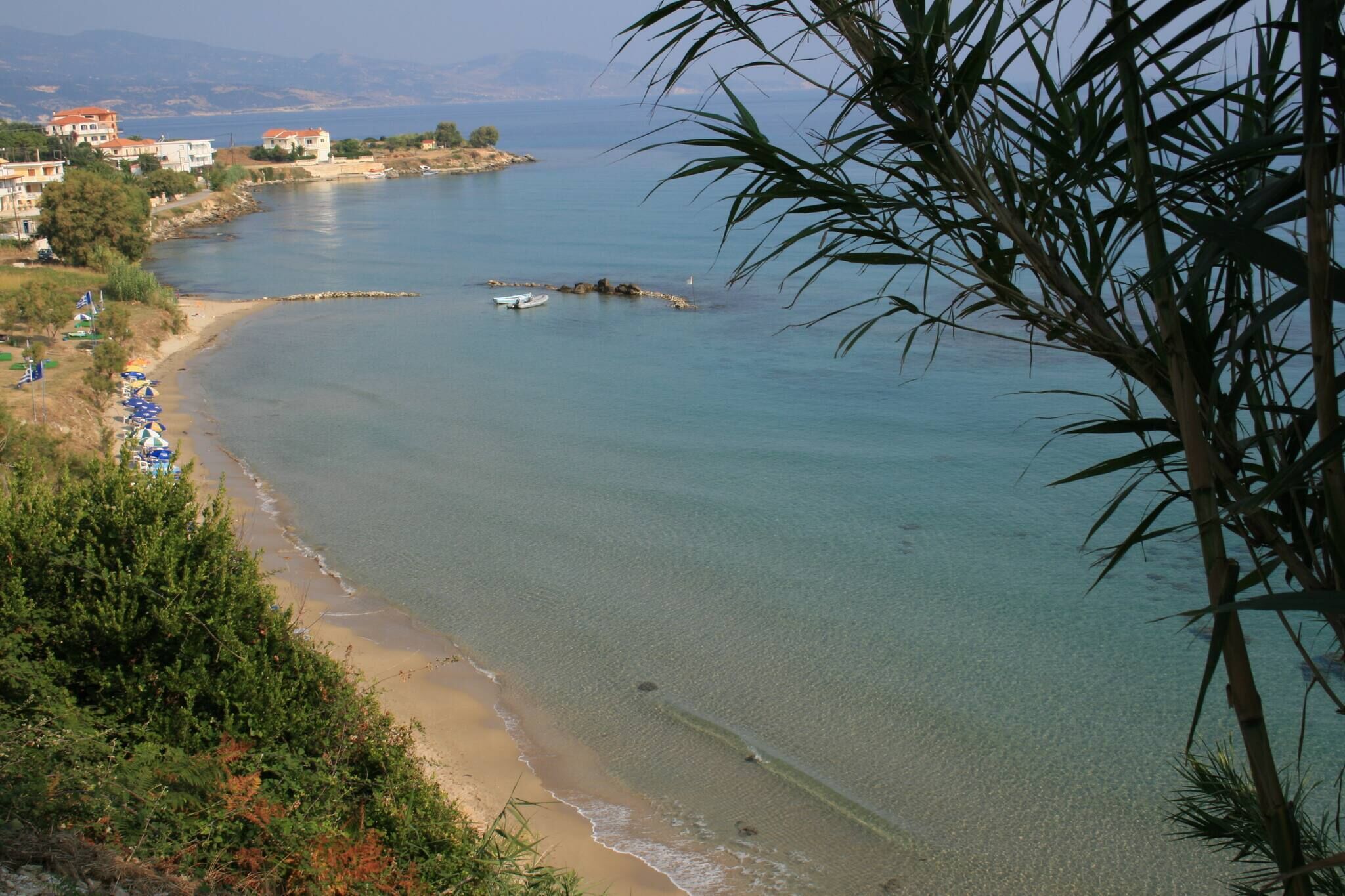 A coastal view with clear water, a sandy beach, and buildings in the background.
