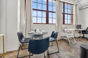 Una habitación con una mesa redonda dispuesta para cuatro personas, dos sillas negras, una silla blanca, una mesita con un ordenador portátil y una ventana con cortinas.