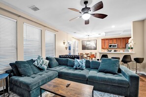 Un salon moderne avec un canapé d’angle, un ventilateur de plafond et un coin cuisine à l’arrière-plan.