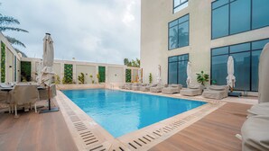 Une piscine entourée de chaises longues et de parasols, elle-même bordée d’un bâtiment doté de grandes fenêtres.