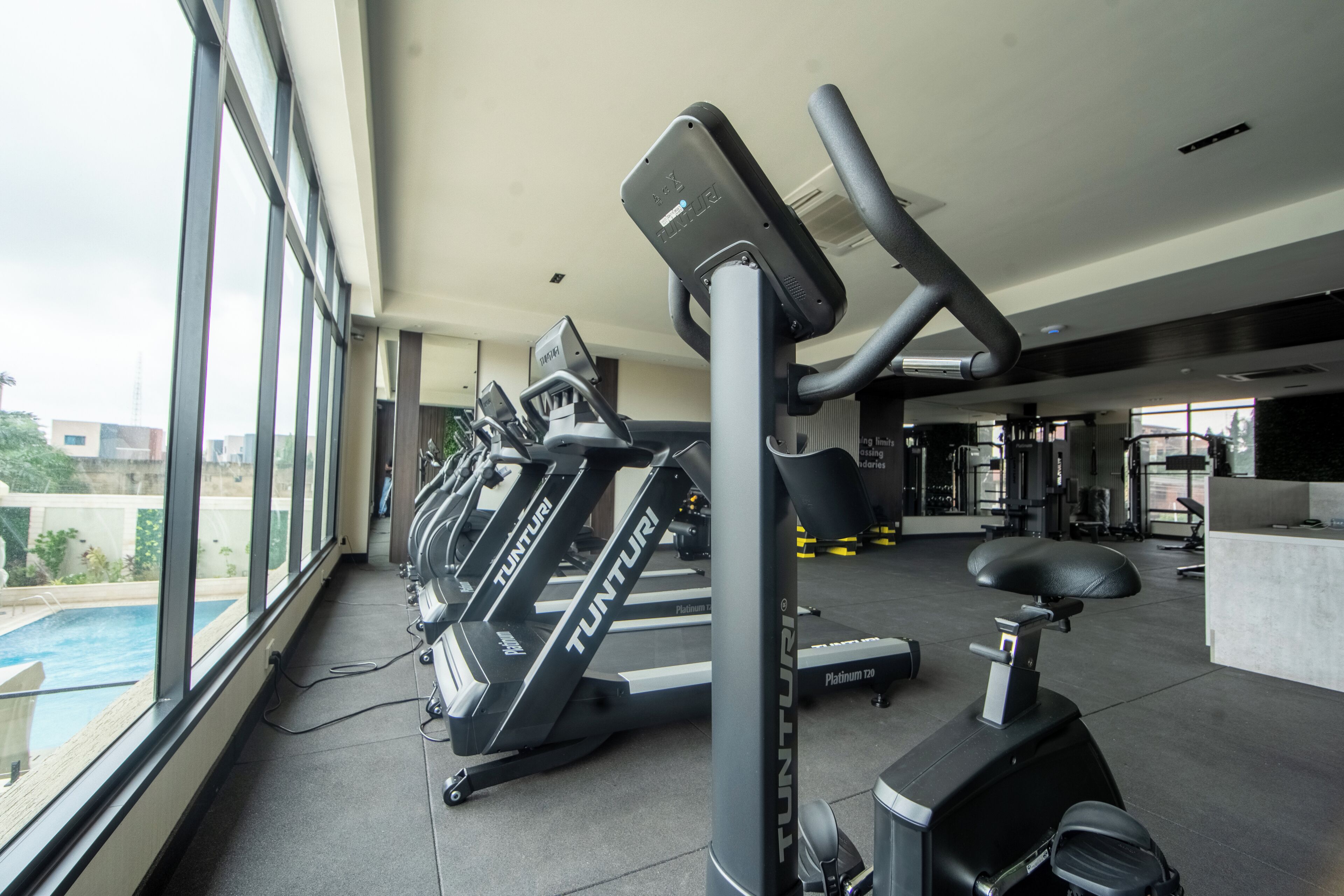 A gym with treadmills, an elliptical, and a rowing machine, with a pool visible through large windows.