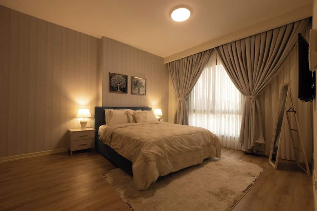 Une chambre à coucher comprenant un lit, des tables de chevet, un tapis, des rideaux et des œuvres d’art murales.
