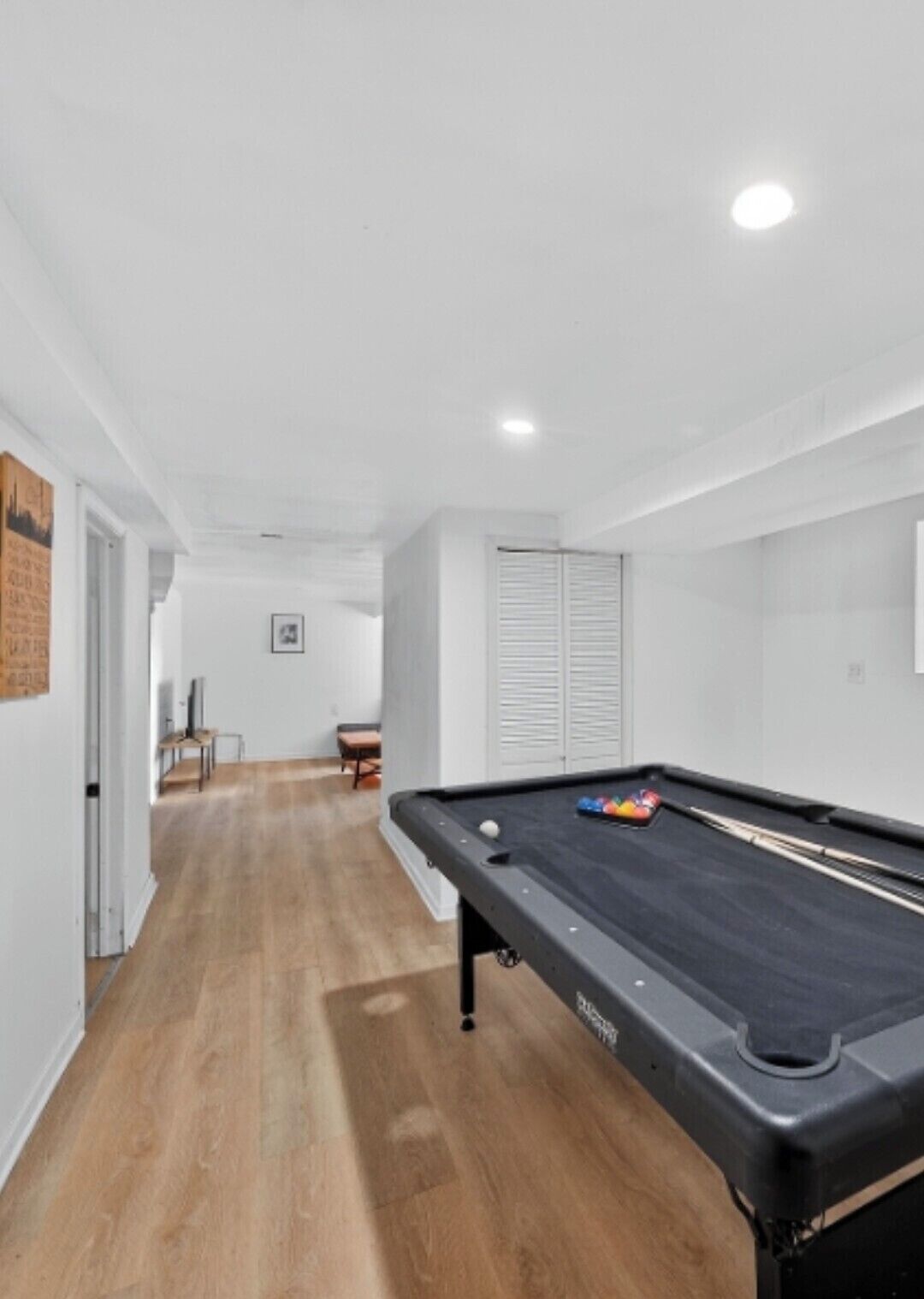 Une table de billard se trouve dans une pièce dont le sol est en bois, les murs sont blancs et une porte donne accès à une autre pièce.