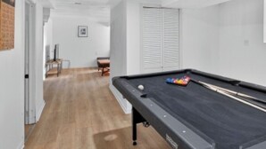 A pool table in a room with wooden flooring, white walls, and a door leading to another room.