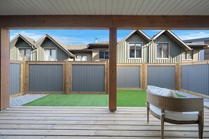 A covered patio with a wooden deck, a wicker chair, and a view of a residential area with houses and a fenced yard. - Brand New Invermere Getaway | Steps to Downtown (Invermere)