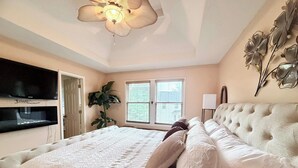 A bedroom with a bed, a television, a ceiling fan, a window with blinds, and a wall-mounted plant.