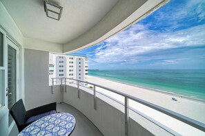 Ein Balkon mit Blick auf einen Strand und das Meer, ein Tisch mit einem gemusterten Tischläufer und ein Gebäude im Hintergrund.