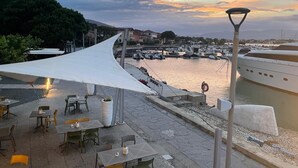 Restaurante à beira-mar com mesas ao ar livre, toldo e vista para barcos e água.