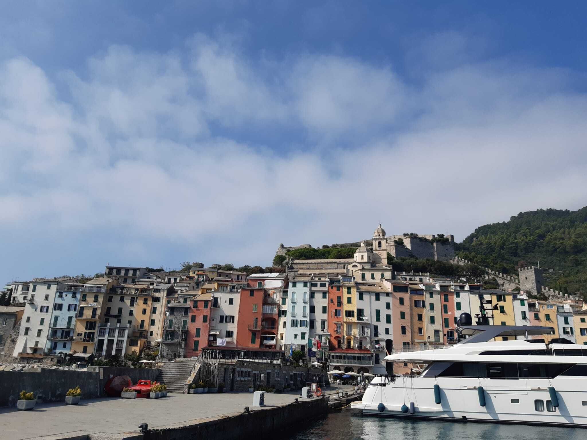 Un marina sul lungomare con edifici colorati, un grande yacht bianco e un'antica fortezza su una collina.