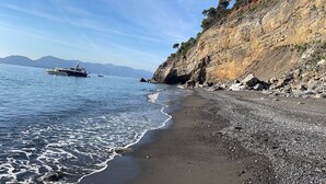Uma praia com costa rochosa, barcos na água e montanhas ao fundo.