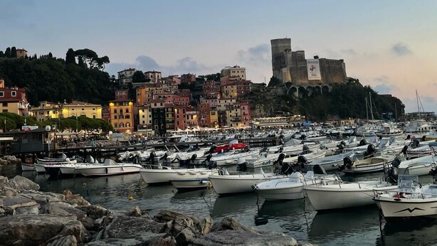 Uma marina com barcos atracados, um edifício histórico ao fundo e terreno rochoso em primeiro plano.