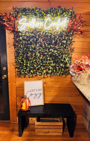 Eine Wand mit grünem Laub im Hintergrund und einem leuchtenden Neon-Schild mit der Aufschrift 'Southern Comfort'. Darunter ein gerahmtes Schild mit der Aufschrift 'Let's Get Cozy' und ein Kerzenhalter mit einer brennenden Kerze.