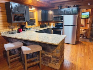 A kitchen with granite countertops, a central island with stools, stainless steel appliances, and wooden cabinetry. - Cozy 3BR Cabin-Bunk Bed Tent Room-1.5mi to Parkway (Pigeon Forge)