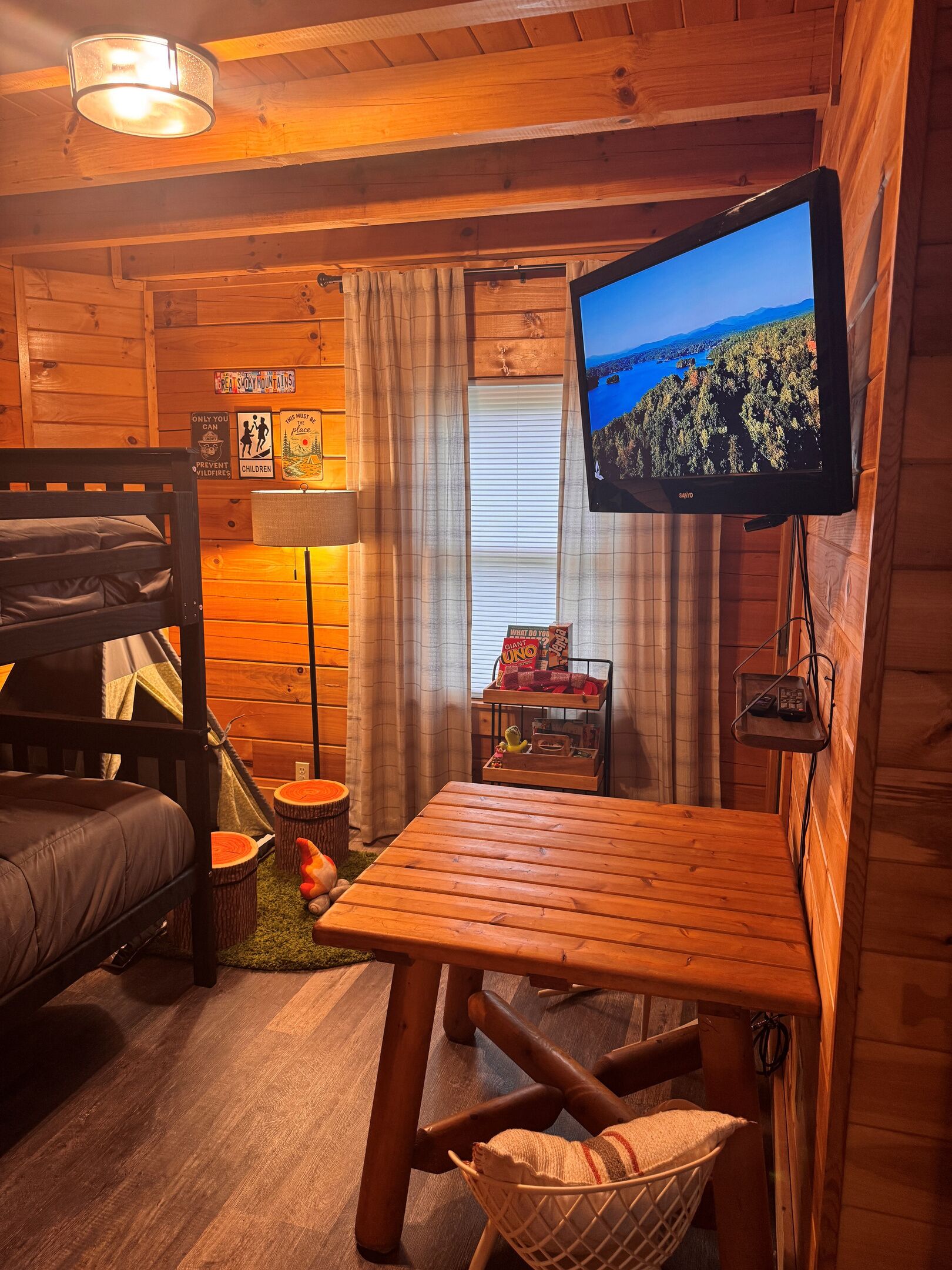 Ein gemütliches Blockhäuschen-Interieur mit einem Etagenbett, einem Holztisch, einem Fernseher und einem Fenster mit Blick auf eine bewaldete Landschaft.
