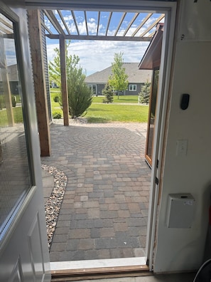 Näkymä oven läpi kivilaatoitetulle terassialueelle, jossa on puinen pergola, nurmikko ja taustalla talo.