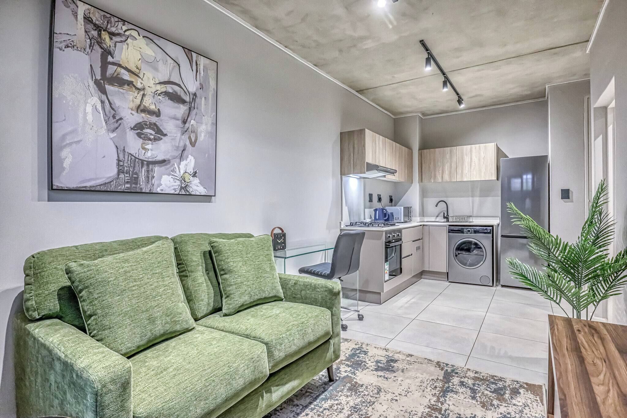 A modern living room with a green sofa, a kitchen with a built-in oven and washing machine, and a large abstract painting on the wall.
