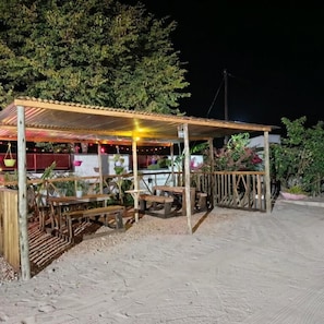 A covered outdoor seating area with wooden benches and tables, surrounded by a wooden fence and plants. - Aroundcorner Guesthouse  (Maun)