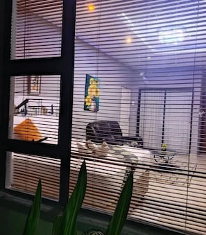 A room with a large window, blinds, a chair, a table, and a plant. - Aroundcorner Guesthouse  (Maun)