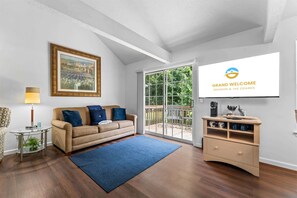 A living room with a sofa, a coffee table, a TV screen displaying 'Grand Welcome Branson & The Ozarks,' and a view through a glass door. - Eagle's View Condo at Fall Creek (Branson)