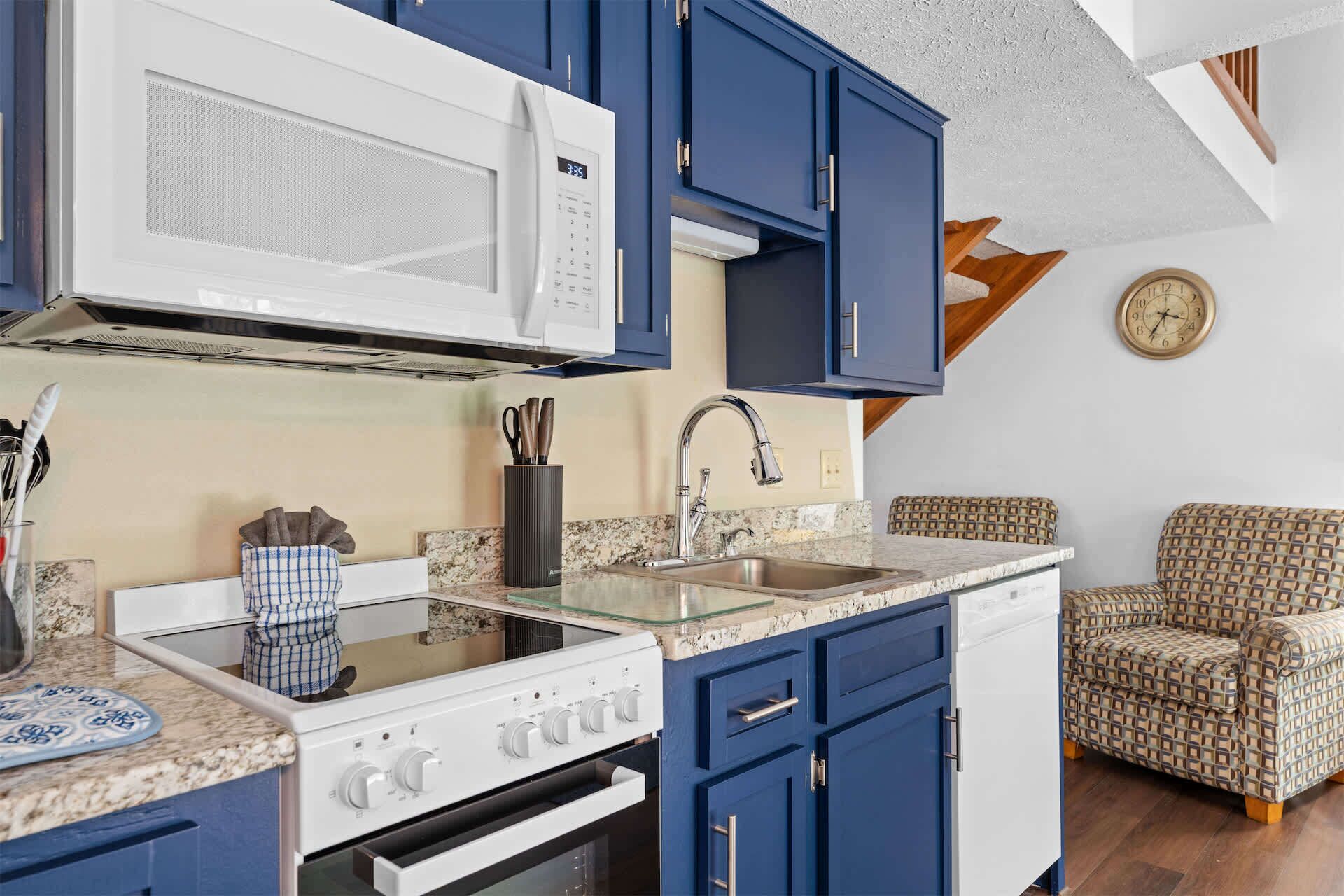A kitchen with a microwave, stove, sink, and blue cabinets. There's a patterned chair and a clock on the wall.