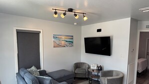 Een woonkamer met een grijze modulaire bank, een grijze fauteuil, een klein rond tafeltje en een flatscreen televisie aan de muur.