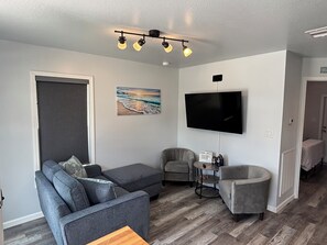 Ein Wohnzimmer mit einem grauen Ecksofa, einem grauen Sessel, einem kleinen runden Tisch und einem an der Wand montierten Flachbildfernseher.