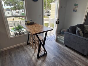 Eine Küche mit einem Holztisch und Hockern, einem grauen Sofa und Blick auf eine Wohngegend durch ein Fenster.