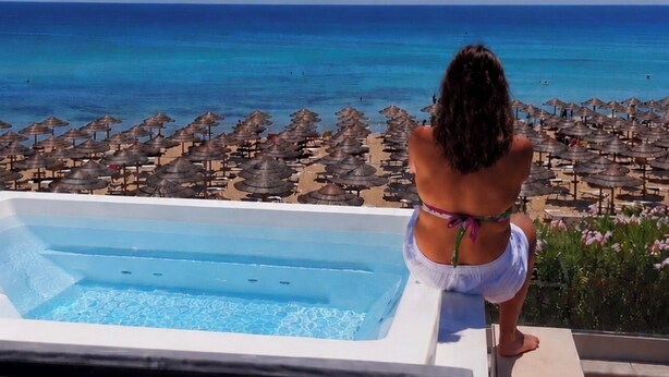 En person i hvid bikini sidder på en liggestol med udsigt over en pool og stranden, med strandparasoller.