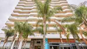 Um hotel com vários andares, varandas, palmeiras e lojas, incluindo uma sanduíche e um restaurante com grelhados.