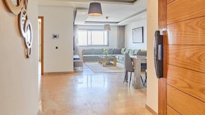Um corredor amplo com porta de madeira, luminária no teto e vista para uma sala de estar com sofá e mesa.