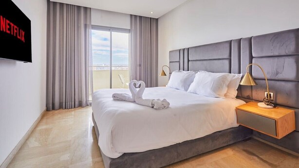 Quarto de hotel com uma cama grande, TV fixada na parede, mesa de cabeceira com abajur e vista do céu pela janela.