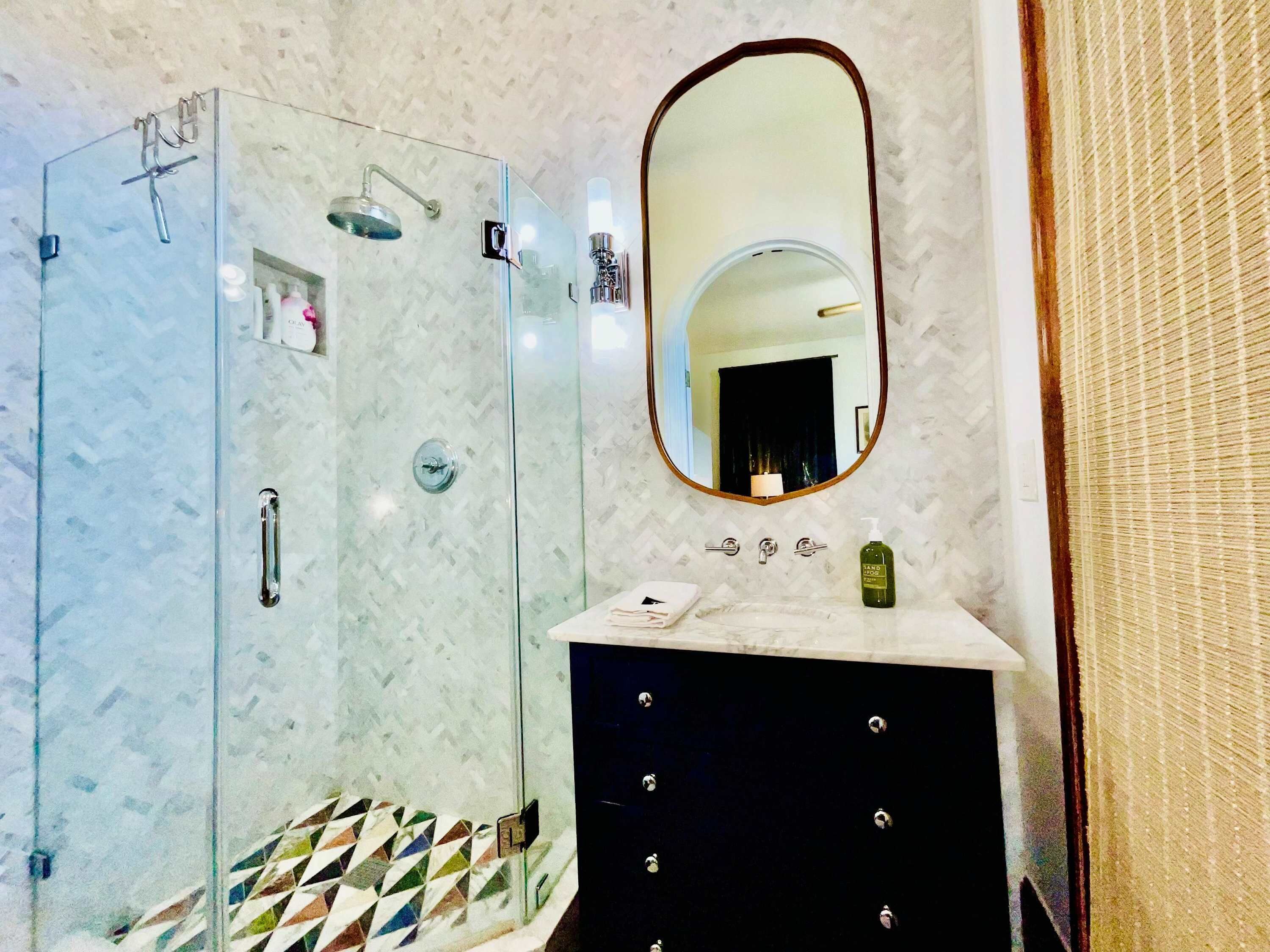 A bathroom with a glass shower enclosure, a vanity with a mirror, and a patterned tile floor.