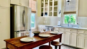 Una cocina con una mesa de madera, taburetes, un refrigerador, armarios y un ventanal con persianas.