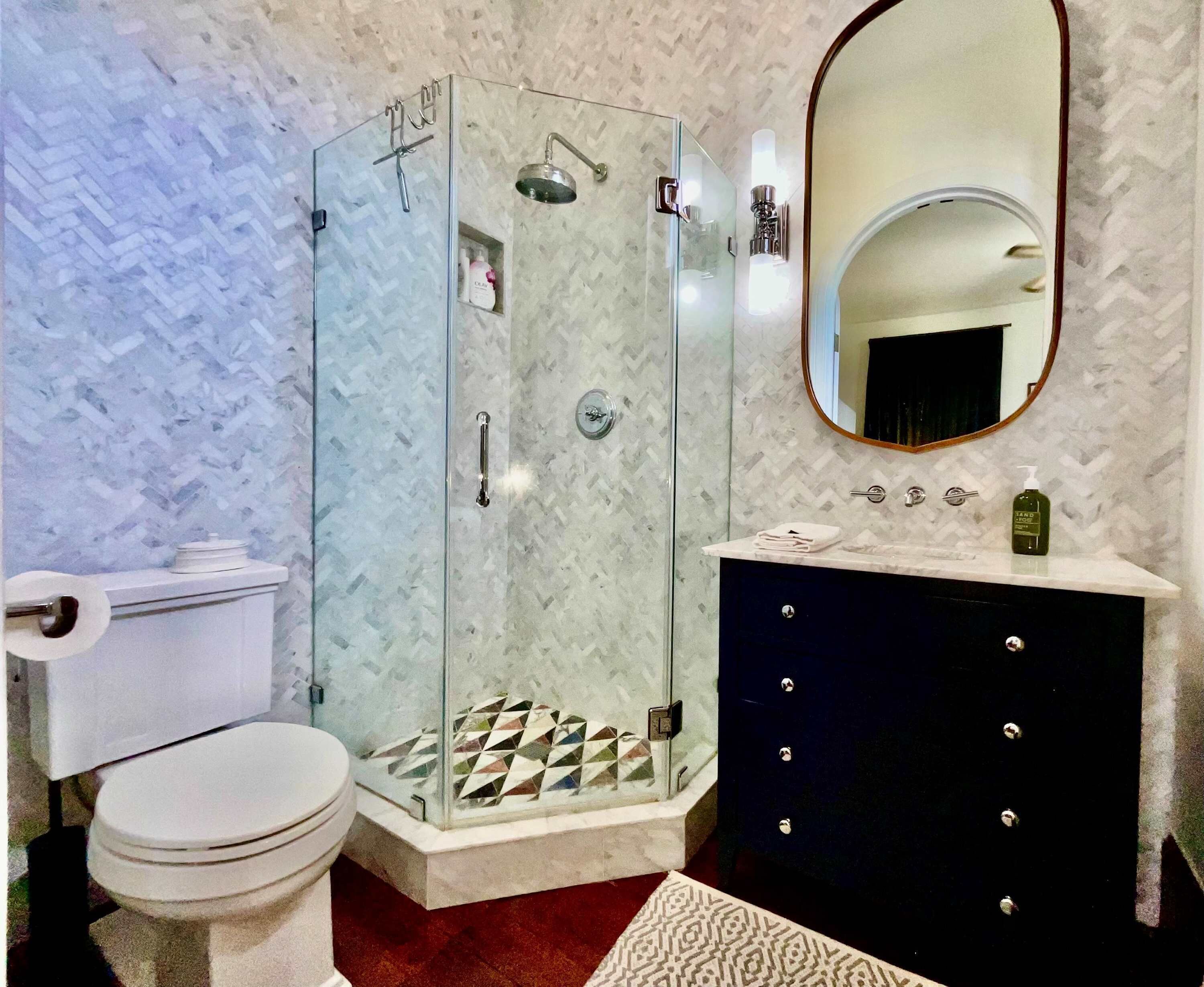 A bathroom with a glass-enclosed shower, a toilet, a vanity with a mirror, and a patterned floor.