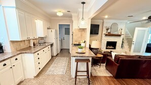 A modern kitchen with white cabinets, a central island, and a dining area with a brown leather sofa. - Refuge on Trinity. Boho Farmhouse, Game Room, Fire Pit (Waxahachie)