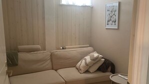A room with a beige sofa, a small round table, a radiator, a window with curtains, and a framed picture on the wall. - Apartment downstairs in my house. Short walking distance to commuter train. (Stockholm)