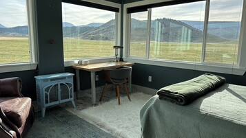 A room with a bed, a desk, a chair, a clock, and large windows with a view of a field and hills.