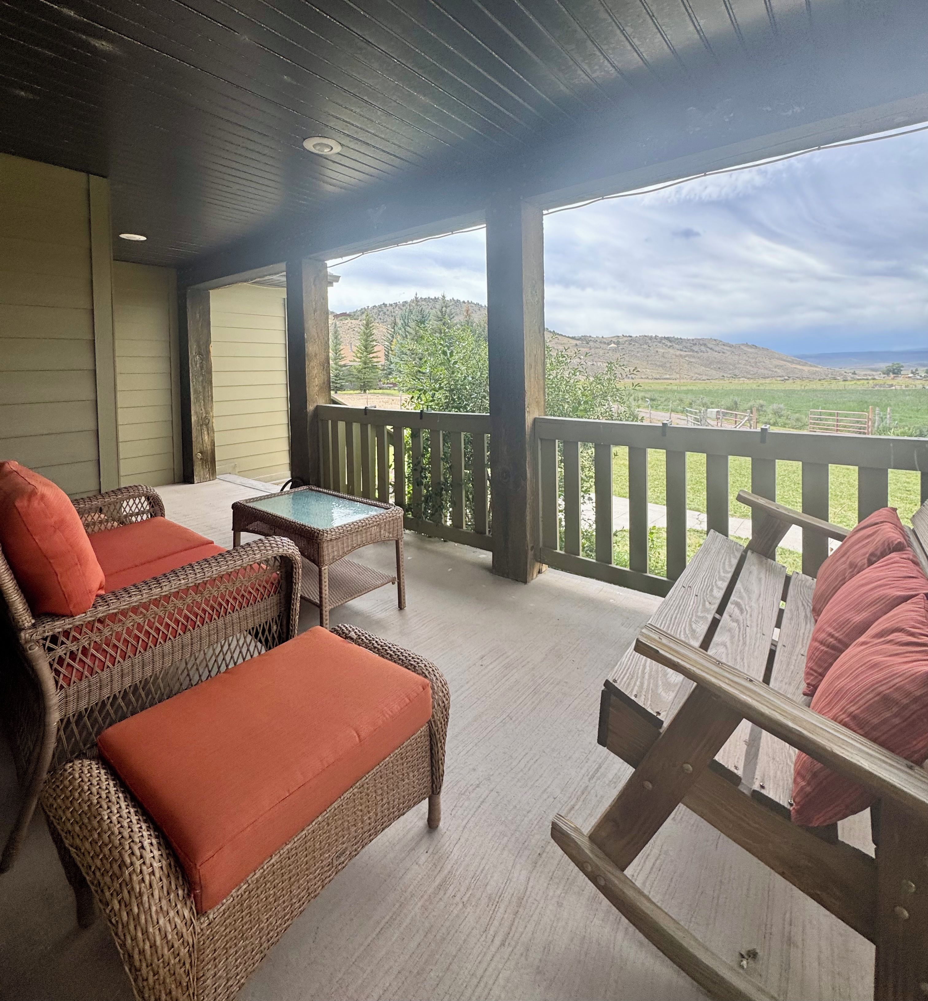 A covered outdoor seating area with wicker furniture and cushions, overlooking a rural landscape.