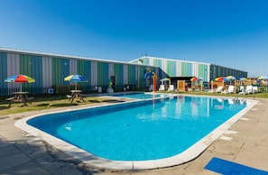 Una piscina con sillas de descanso y sombrillas, un edificio con revestimiento vertical y un cielo azul despejado.