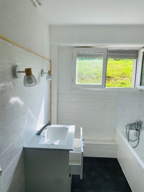 Et badeværelse med hvid vask, spejl, et vindue med udsigt til grøn natur og et badekar.