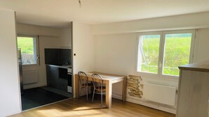 Quarto com mesa de jantar de madeira, duas cadeiras, área de cozinha com armários e fogão, e janela com vista para vegetação.