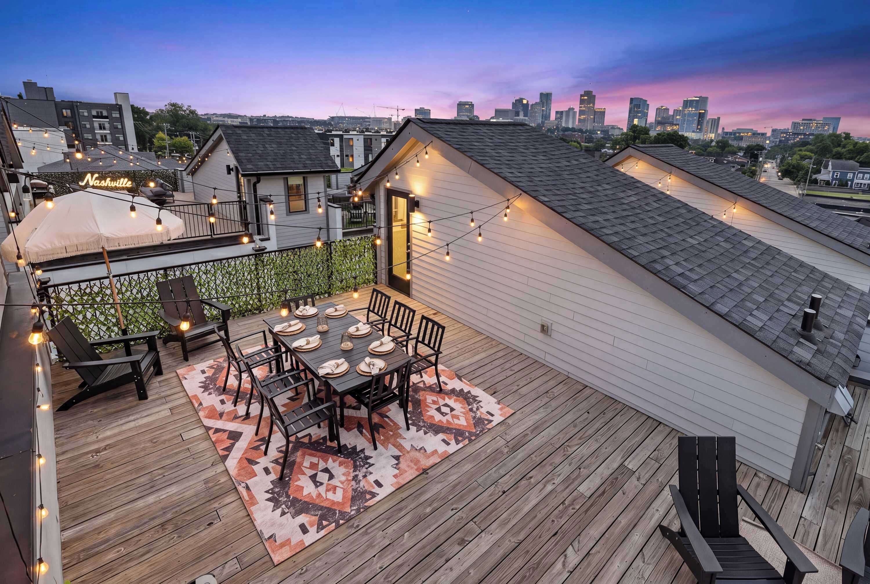A rooftop patio with a dining table, chairs, string lights, and a city skyline view.