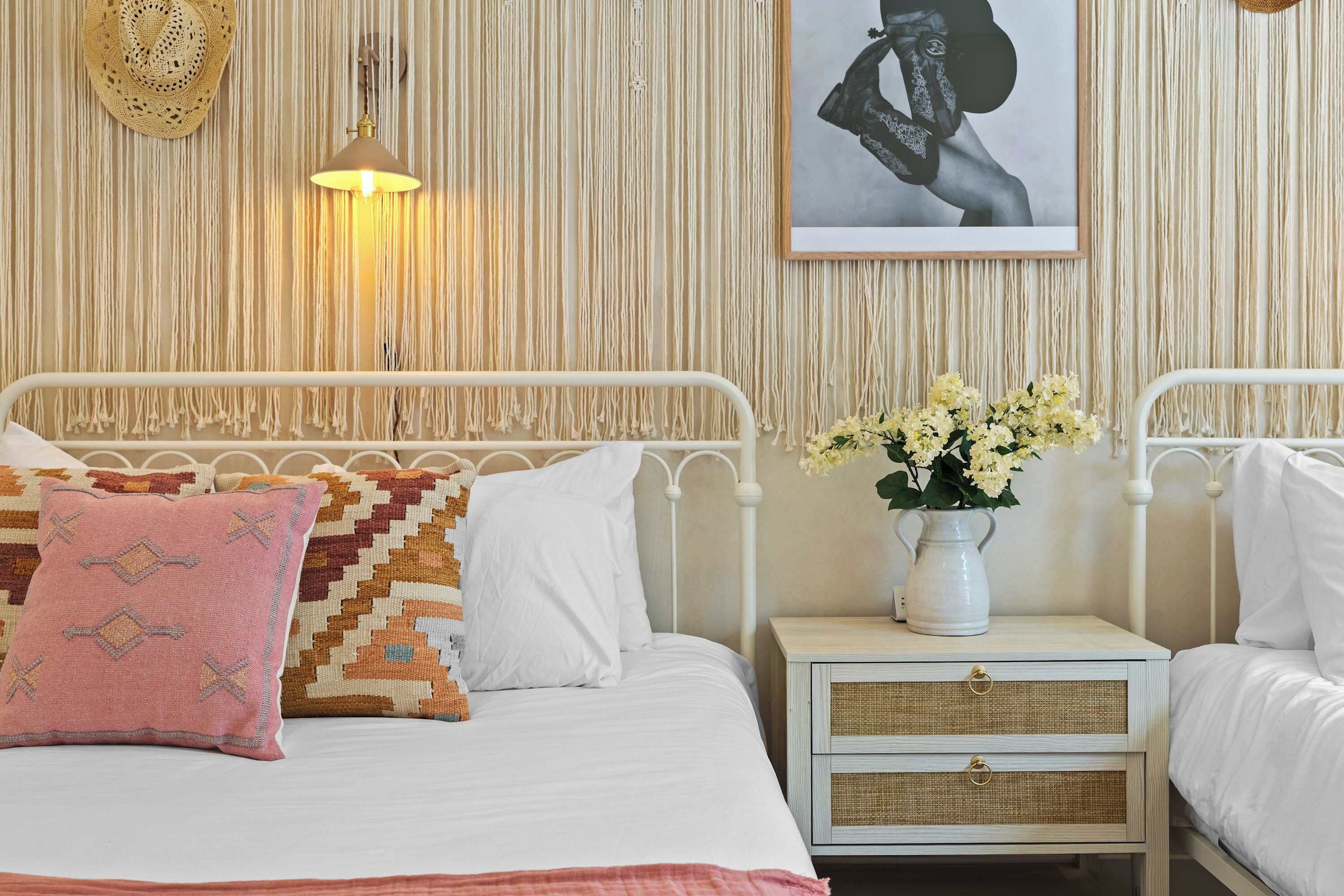 A bed with white bedding, a patterned pillow, a bedside table with a vase of flowers, and a wall with a macramé wall hanging and a framed picture.
