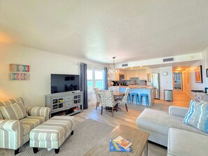 A modern living room with a TV, sofa, ottoman, and a coffee table with a magazine. - Vacation Villas #635 Renovated 6th Floor Beachfront Condo (Fort Myers Beach)