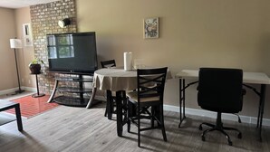 Ein Wohnzimmer mit Fernseher, einem runden Tisch, zwei Stühlen, einem kleinen Tisch mit Lampe, einer Topfpflanze und einer Ziegelwand.