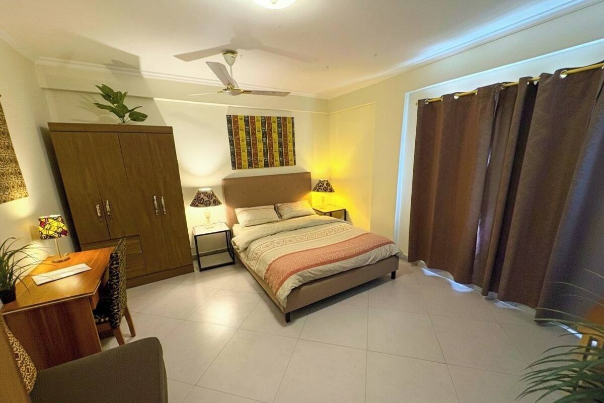 A bedroom with a bed, a desk, a chair, a wardrobe, a ceiling fan, and a window with curtains.