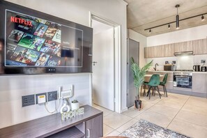 Ein modernes Wohnzimmer mit einem großen Flachbildfernseher, der Inhalte von Netflix zeigt, eine minimalistische Küche mit Edelstahlgeräten und ein Essbereich mit grünen Stühlen.