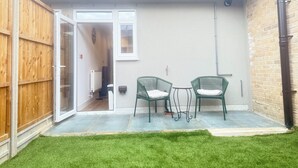 Une terrasse avec deux chaises vertes et une petite table, une clôture en bois et l’entrée d’un bâtiment.